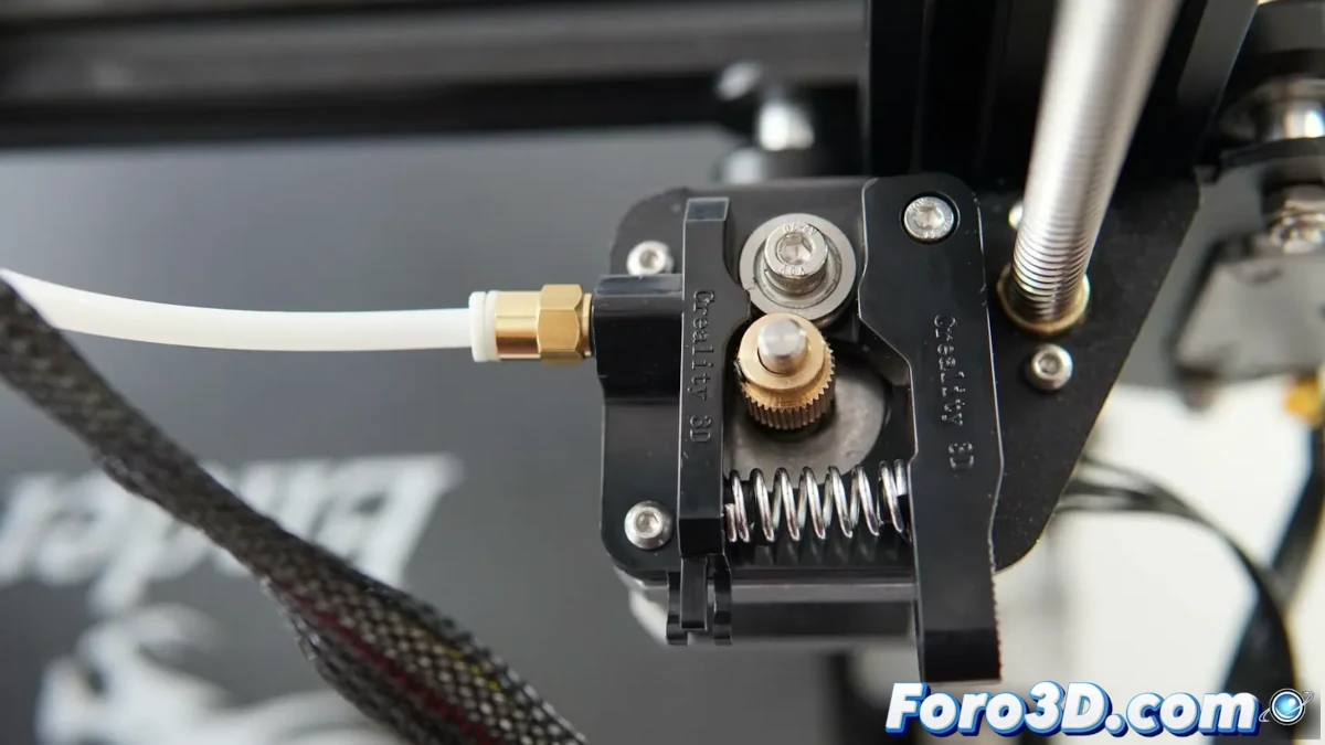 Un primer plano del extrusor de una impresora 3D Creality Ender 3 con modificaciones específicas para imprimir filamento TPU. Se muestra un extrusor metálico, un hotend mejorado y un tubo Bowden optimizado. La imagen incluye una bobina de TPU flexible y una impresión en proceso con detalles visibles en la base de impresión.